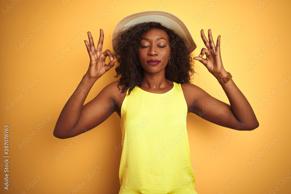 Young african american woman wearing t-shirt and hat over isolated yellow background relax and smiling with eyes closed doing meditation gesture with fingers. Yoga concept. Young african american woman wearing t-shirt and hat over isolated yellow background relax and smiling with eyes closed doing meditation gesture with fingers. Yoga concept.