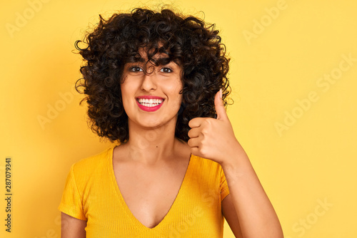 Young arab woman with curly hair wearing t-shirt standing over isolated yellow background happy with big smile doing ok sign, thumb up with fingers, excellent sign