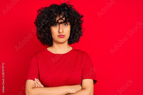 Young arab woman with curly hair wearing casual t-shirt over isolated red background skeptic and nervous, disapproving expression on face with crossed arms. Negative person.