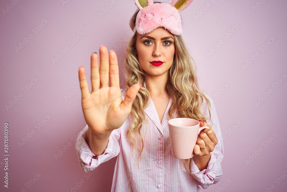 Young woman wearing pajama and mask drinking a cup of coffee over pink isolated background with open hand doing stop sign with serious and confident expression, defense gesture Young woman wearing pajama and mask drinking a cup of coffee over pink isolated background with open hand doing stop sign with serious and confident expression, defense gesture