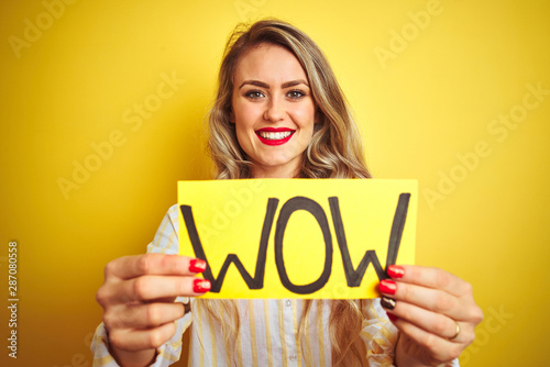 Beautiful woman holding amazed wow surprise banner over isolated yellow background with a happy face standing and smiling with a confident smile showing teeth