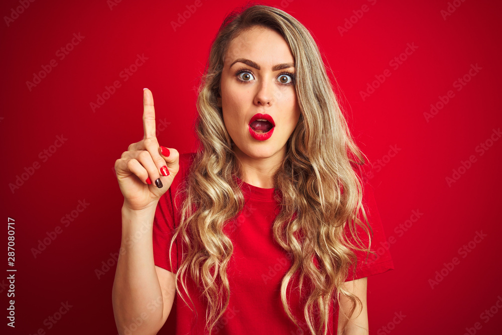 Young beautiful woman wearing basic t-shirt standing over red isolated background pointing finger up with successful idea. Exited and happy. Number one. Young beautiful woman wearing basic t-shirt standing over red isolated background pointing finger up with successful idea. Exited and happy. Number one.