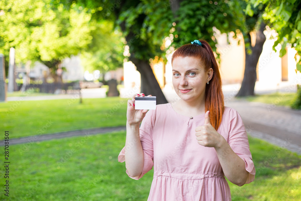 Young caucasian woman stay in park and show white credit card with black magnet line. Shopping and retail concept. Plastic bank-card design mock up Young caucasian woman stay in park and show white credit card with black magnet line. Shopping and retail concept. Plastic bank-card design mock up