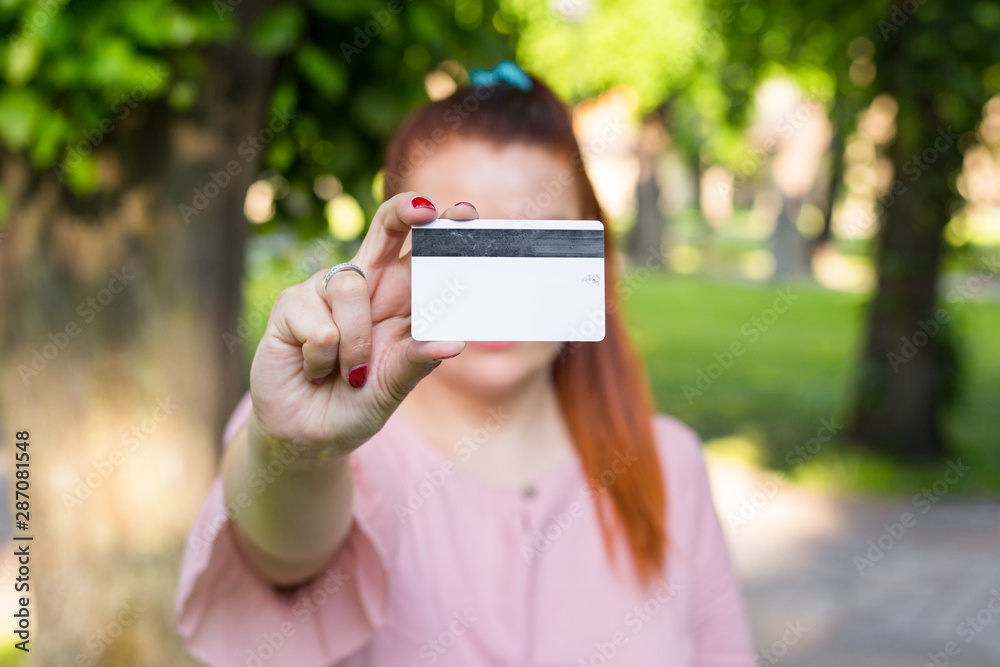 Young caucasian woman stay in park and show white credit card with black magnet line. Shopping and retail concept. Plastic bank-card design mock up