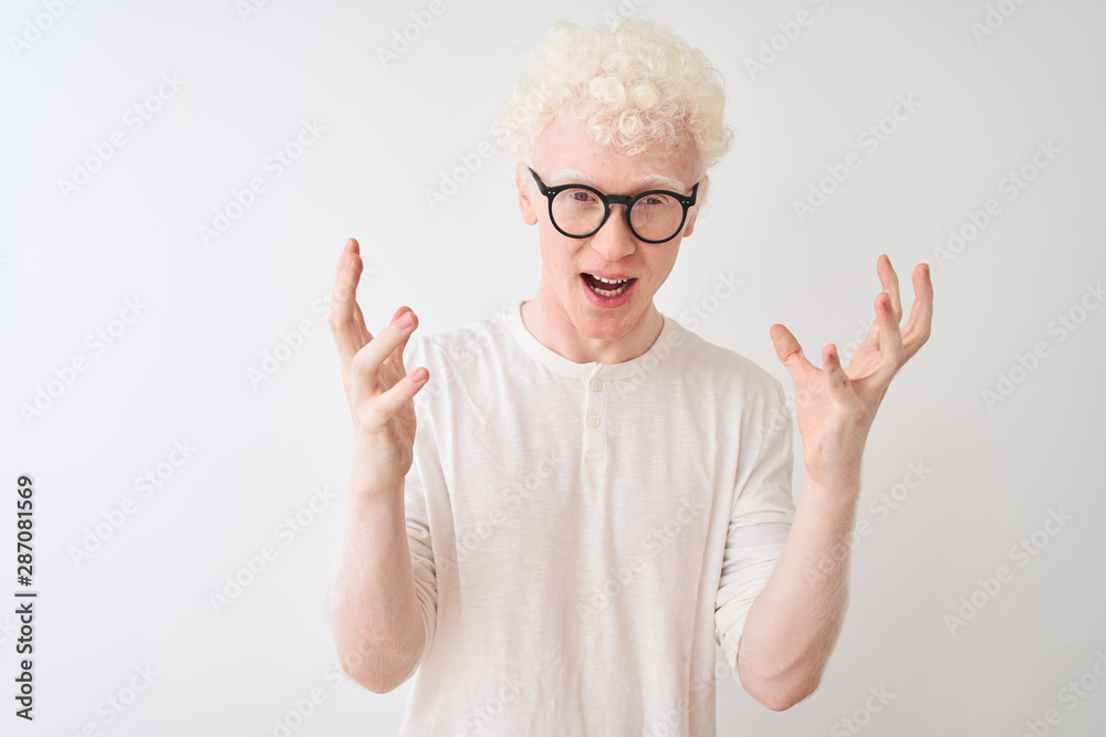 Young albino blond man wearing t-shirt and glasses standing over isolated white background celebrating crazy and amazed for success with arms raised and open eyes screaming excited. Winner concept