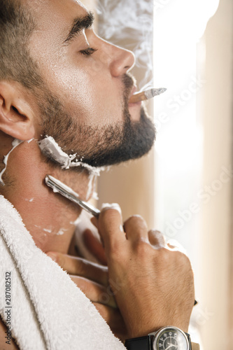 Man smoking a cigarette and shaving his beard with a straight razor