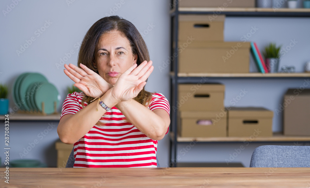 Middle age senior woman sitting at the table at home Rejection expression crossing arms and palms doing negative sign, angry face Middle age senior woman sitting at the table at home Rejection expression crossing arms and palms doing negative sign, angry face