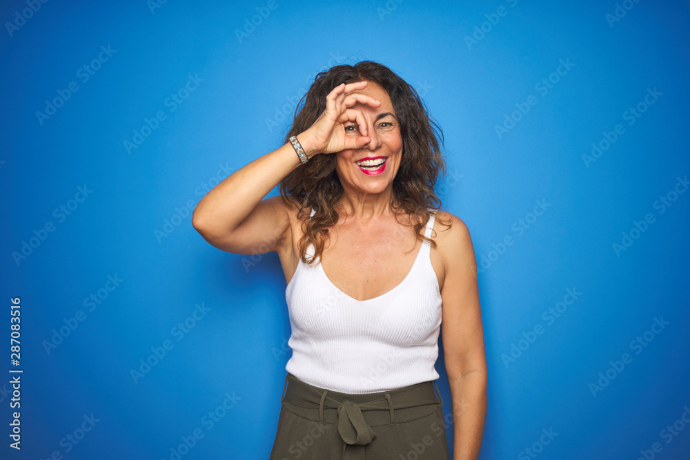 Middle age senior woman with curly hair standing over blue isolated background doing ok gesture with hand smiling, eye looking through fingers with happy face. Middle age senior woman with curly hair standing over blue isolated background doing ok gesture with hand smiling, eye looking through fingers with happy face.