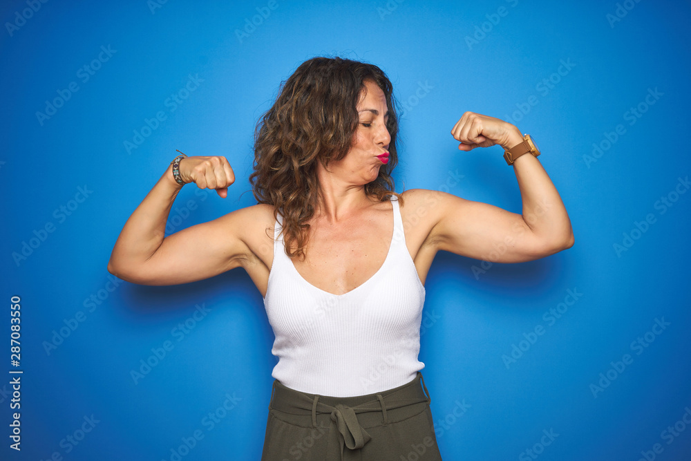 Middle age senior woman with curly hair standing over blue isolated background showing arms muscles smiling proud. Fitness concept. Middle age senior woman with curly hair standing over blue isolated background showing arms muscles smiling proud. Fitness concept.