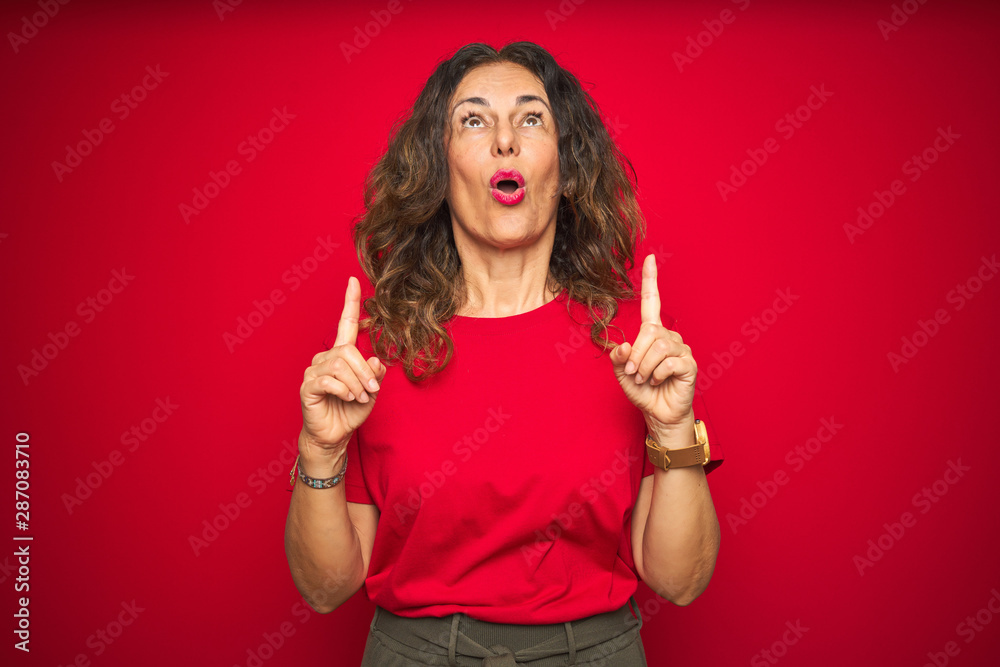 Middle age senior woman with curly hair over red isolated background amazed and surprised looking up and pointing with fingers and raised arms. Middle age senior woman with curly hair over red isolated background amazed and surprised looking up and pointing with fingers and raised arms.