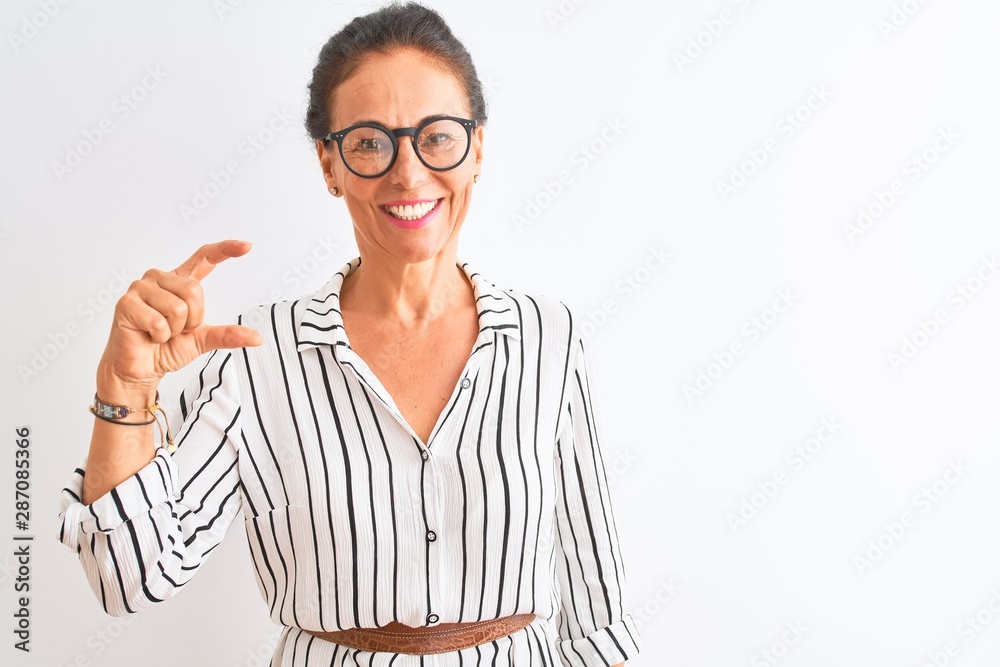 Middle age businesswoman wearing striped dress and glasses over isolated white background smiling and confident gesturing with hand doing small size sign with fingers looking and the camera