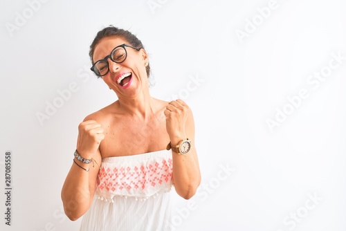 Middle age woman wearing casual dress and glasses standing over isolated white background very happy and excited doing winner gesture with arms raised, smiling and screaming for success. Celebration
