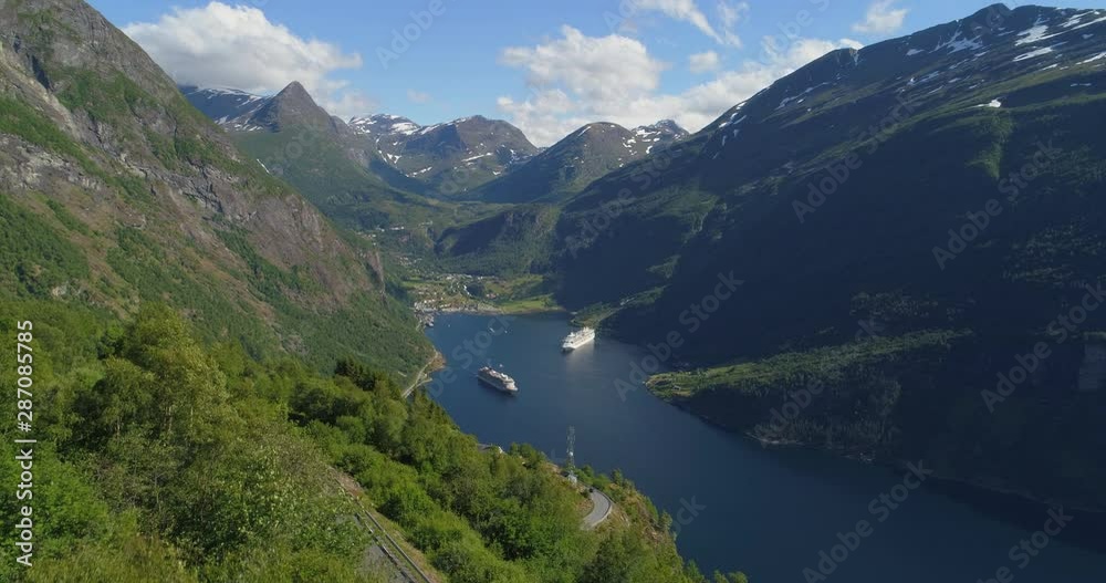 Aerial: Drone approaching ships in Geirangerfjord amidst mountains with winding road on sunny day during winter - Geiranger Fjord, Norway