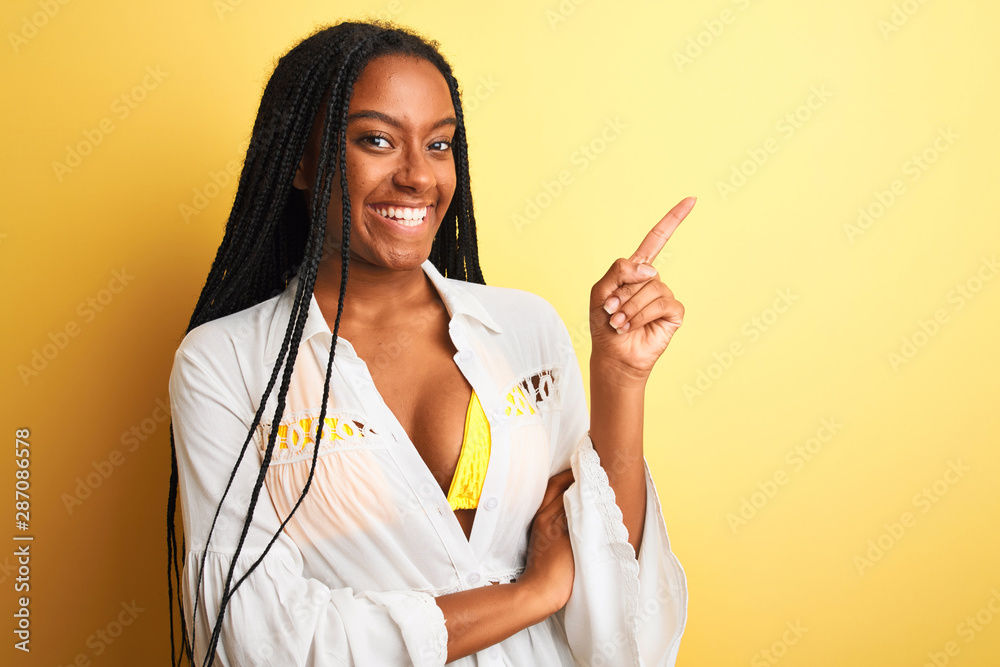 African american woman on vacation wearing bikini and shirt over isolated yellow background with a big smile on face, pointing with hand and finger to the side looking at the camera. African american woman on vacation wearing bikini and shirt over isolated yellow background with a big smile on face, pointing with hand and finger to the side looking at the camera.