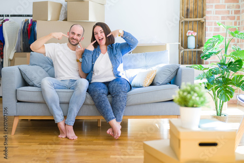 Young couple sitting on the sofa arround cardboard boxes moving to a new house smiling cheerful showing and pointing with fingers teeth and mouth. Dental health concept. © Krakenimages.com