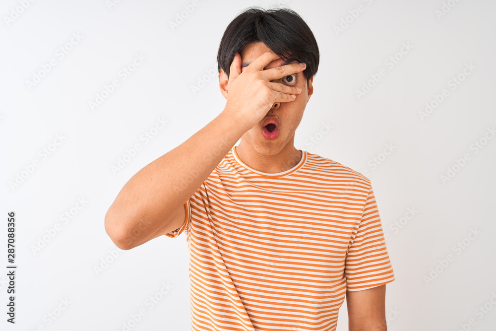 Young chinese man wearing casual striped t-shirt standing over isolated white background peeking in shock covering face and eyes with hand, looking through fingers with embarrassed expression. Young chinese man wearing casual striped t-shirt standing over isolated white background peeking in shock covering face and eyes with hand, looking through fingers with embarrassed expression.