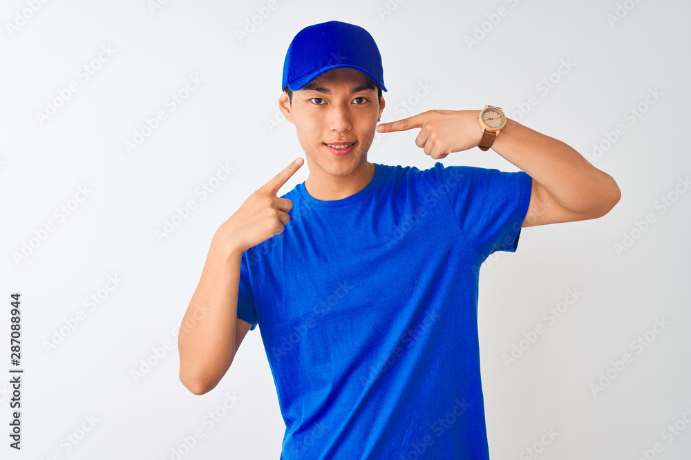 Chinese deliveryman wearing blue t-shirt and cap standing over isolated white background smiling cheerful showing and pointing with fingers teeth and mouth. Dental health concept.