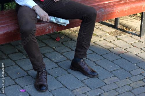 A stylishly dressed young man sits on a park bench and holds a newspaper in his hands. Close up visible legs.