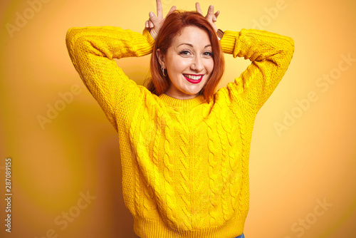 Beautiful redhead woman wearing winter sweater standing over isolated yellow background Posing funny and crazy with fingers on head as bunny ears, smiling cheerful © Krakenimages.com