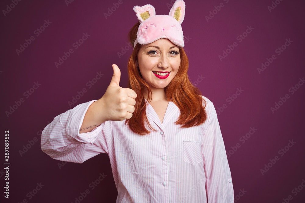 Young beautiful redhead woman wearing pajama and mask over purple isolated background doing happy thumbs up gesture with hand. Approving expression looking at the camera with showing success. Young beautiful redhead woman wearing pajama and mask over purple isolated background doing happy thumbs up gesture with hand. Approving expression looking at the camera with showing success.