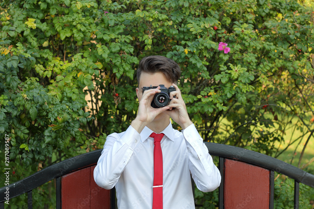 Stylishly dressed young man sits on a park bench and takes pictures. In his hands holds a SLR film camera. Stylishly dressed young man sits on a park bench and takes pictures. In his hands holds a SLR film camera.