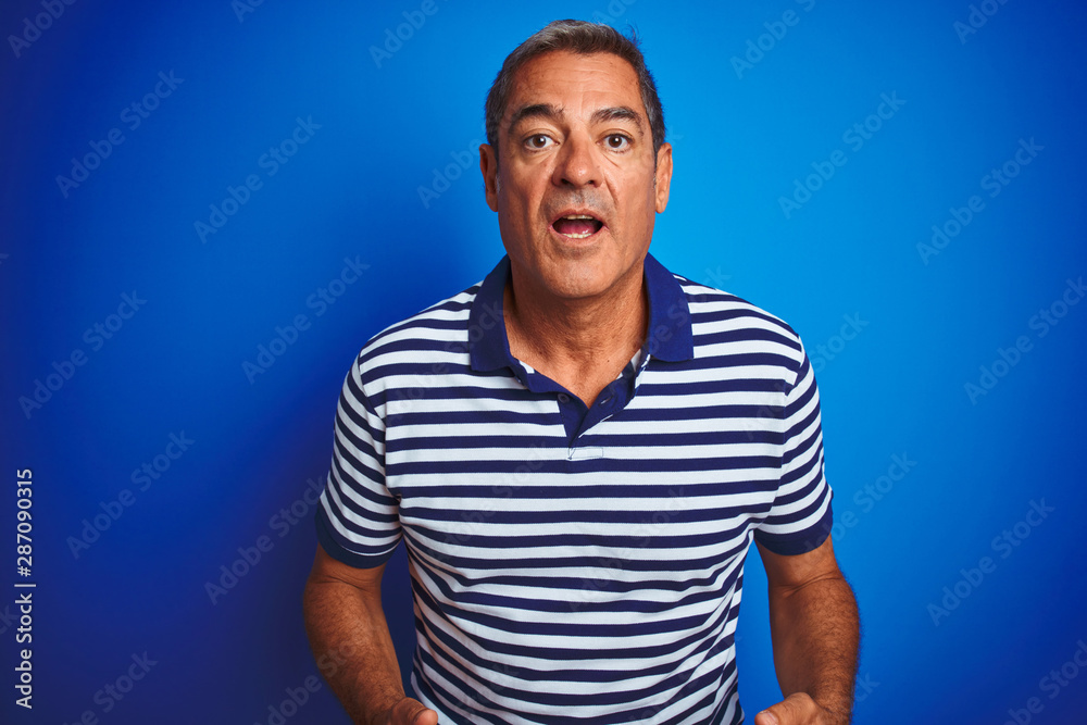 Handsome middle age man wearing striped polo standing over isolated blue background afraid and shocked with surprise expression, fear and excited face. Handsome middle age man wearing striped polo standing over isolated blue background afraid and shocked with surprise expression, fear and excited face.