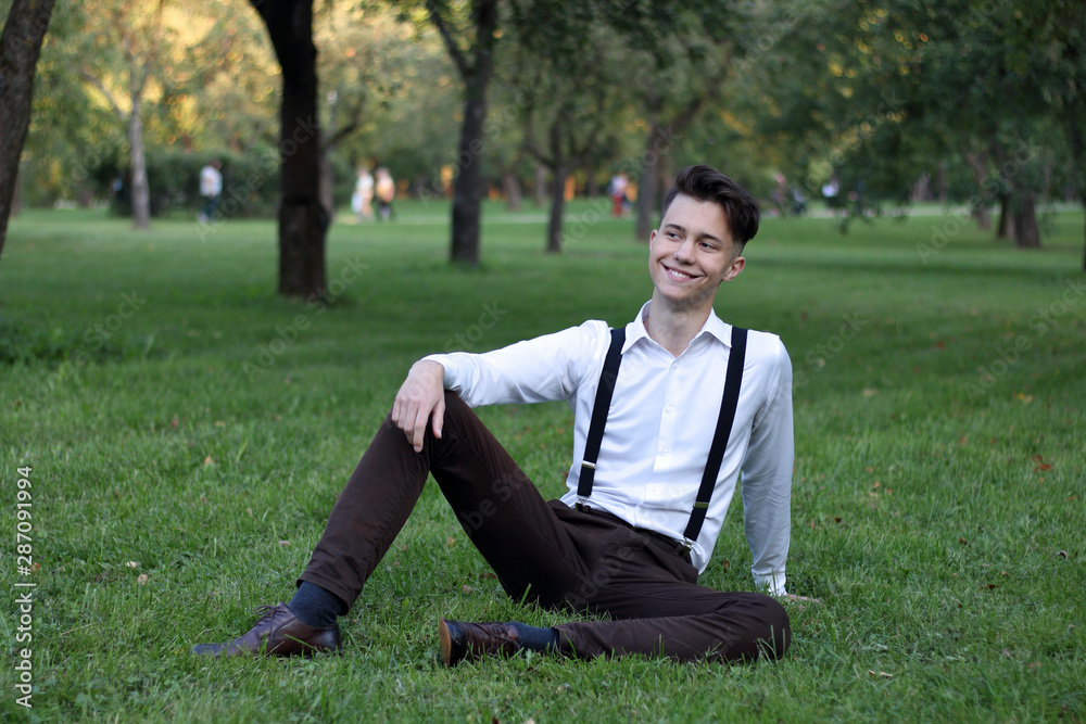 Stylishly dressed young man posing in a park. Sits on the lawn and looks away with a smile. Stylishly dressed young man posing in a park. Sits on the lawn and looks away with a smile.