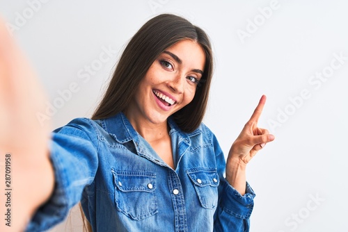 Beautiful woman wearing denim shirt make selfie by camera over isolated white background with a big smile on face, pointing with hand finger to the side looking at the camera.