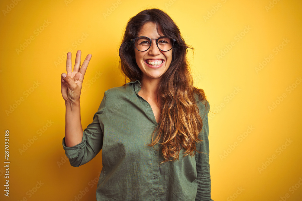Young beautiful woman wearing green shirt and glasses over yelllow isolated background showing and pointing up with fingers number three while smiling confident and happy. Young beautiful woman wearing green shirt and glasses over yelllow isolated background showing and pointing up with fingers number three while smiling confident and happy.
