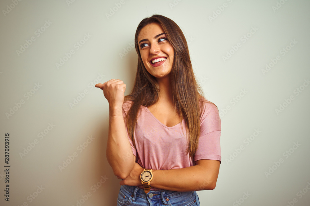 Young beautiful woman wearing casual t-shirt standing over isolated white background smiling with happy face looking and pointing to the side with thumb up. Young beautiful woman wearing casual t-shirt standing over isolated white background smiling with happy face looking and pointing to the side with thumb up.