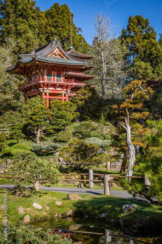 Majestic red japanese house sits on top of a hill with perfect shrubs, trees, water and paths