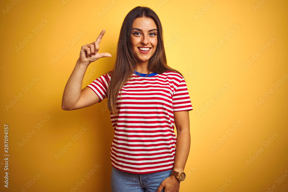 Young beautiful woman wearing striped t-shirt standing over isolated yellow background smiling and confident gesturing with hand doing small size sign with fingers looking and the camera Young beautiful woman wearing striped t-shirt standing over isolated yellow background smiling and confident gesturing with hand doing small size sign with fingers looking and the camera