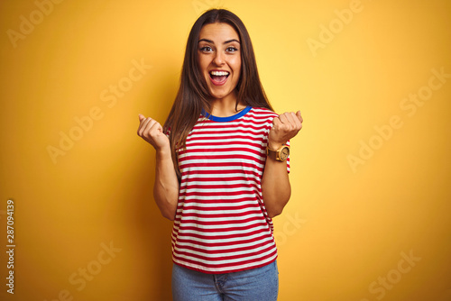 Young beautiful woman wearing striped t-shirt standing over isolated yellow background celebrating surprised and amazed for success with arms raised and open eyes. Winner concept.