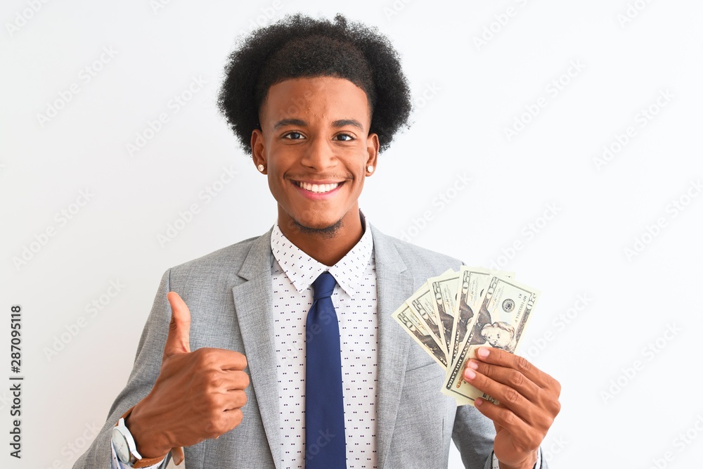 Young african american businessman holding dollars standing over isolated white background happy with big smile doing ok sign, thumb up with fingers, excellent sign Young african american businessman holding dollars standing over isolated white background happy with big smile doing ok sign, thumb up with fingers, excellent sign