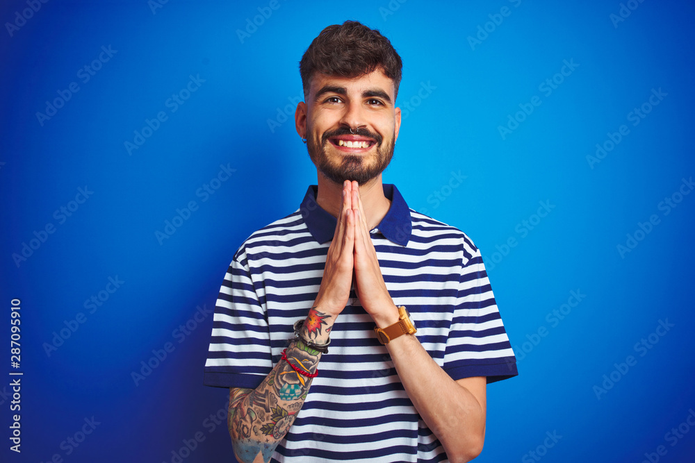 Young man with tattoo wearing striped polo standing over isolated blue background praying with hands together asking for forgiveness smiling confident.