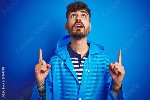 Young handsome man wearing rain coat standing over isolated blue background amazed and surprised looking up and pointing with fingers and raised arms.