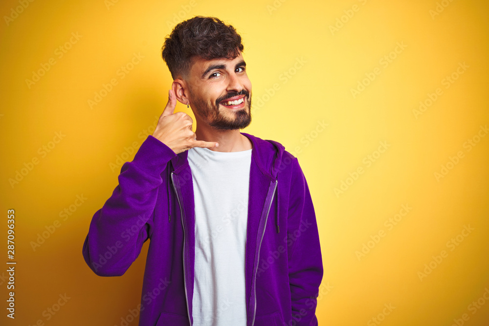 Young man with tattoo wearing sport purple sweatshirt over isolated yellow background smiling doing phone gesture with hand and fingers like talking on the telephone. Communicating concepts. Young man with tattoo wearing sport purple sweatshirt over isolated yellow background smiling doing phone gesture with hand and fingers like talking on the telephone. Communicating concepts.