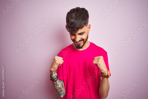 Young man with tattoo wearing t-shirt standing over isolated pink background very happy and excited doing winner gesture with arms raised, smiling and screaming for success. Celebration concept.