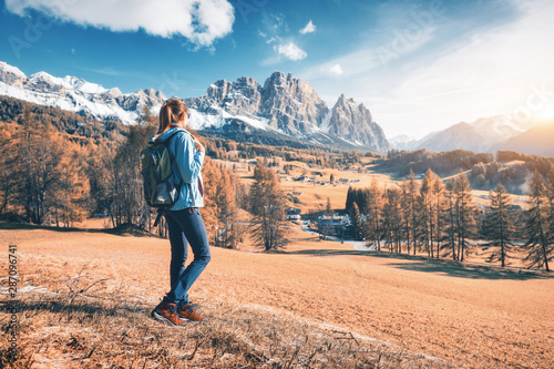 Beautiful young woman with backpack is standing on the hill against mountains at sunset in autumn. Landscape with sporty girl, rocks with snowy peaks, meadow, orange trees, blue sky in Italy. Travel
