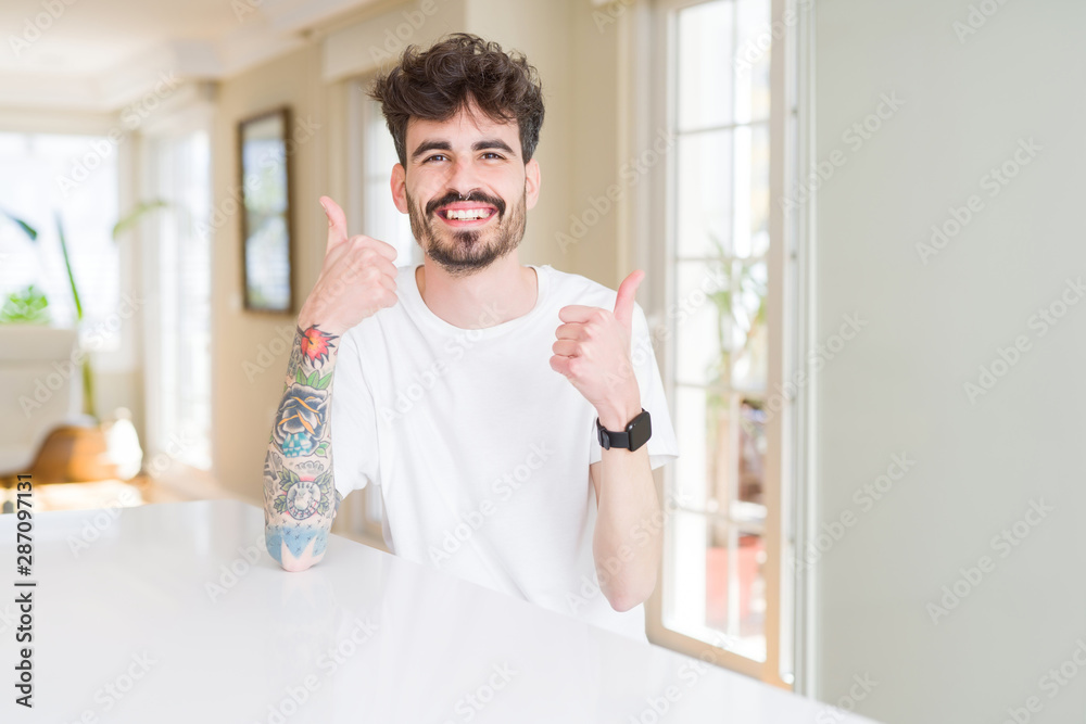 Young man wearing casual t-shirt sitting on white table success sign doing positive gesture with hand, thumbs up smiling and happy. Looking at the camera with cheerful expression, winner gesture. Young man wearing casual t-shirt sitting on white table success sign doing positive gesture with hand, thumbs up smiling and happy. Looking at the camera with cheerful expression, winner gesture.