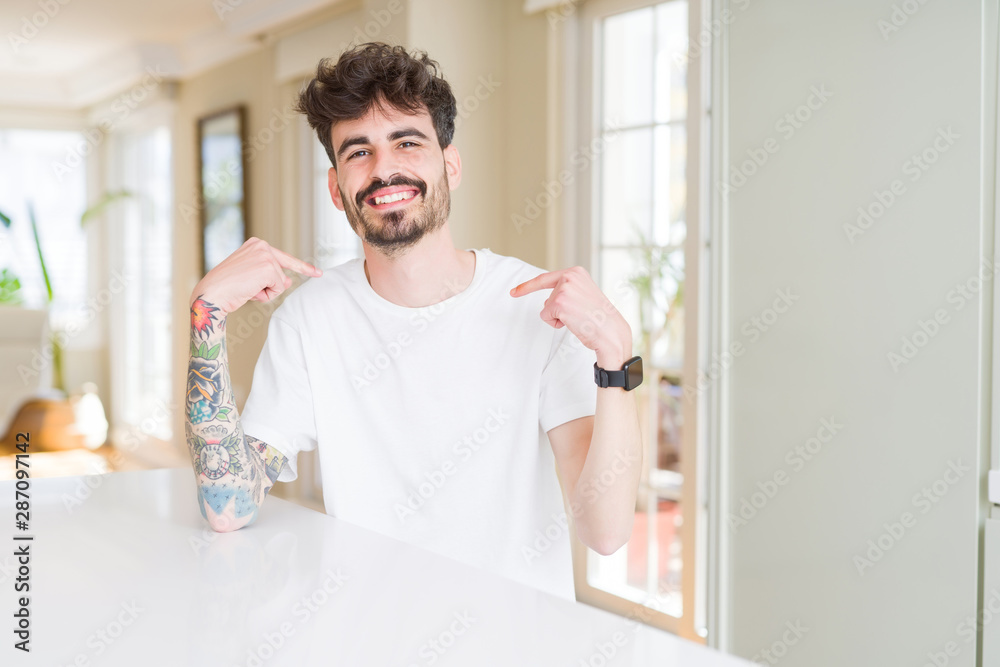 Young man wearing casual t-shirt sitting on white table looking confident with smile on face, pointing oneself with fingers proud and happy.