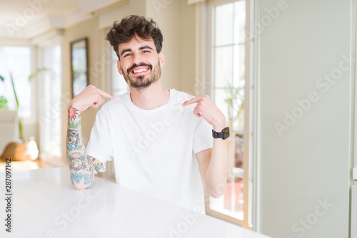Young man wearing casual t-shirt sitting on white table looking confident with smile on face, pointing oneself with fingers proud and happy.