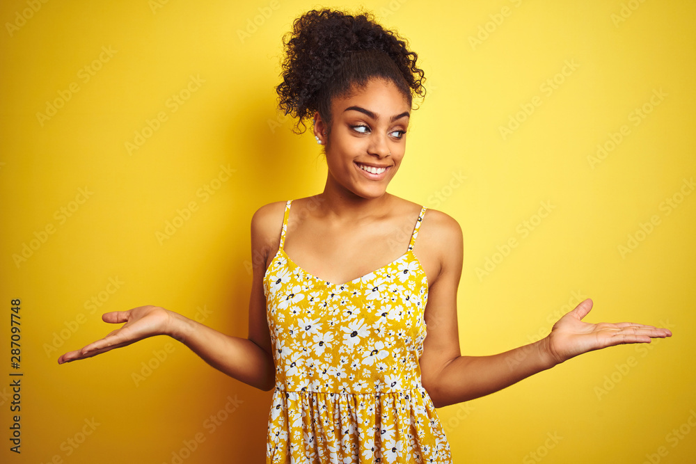 African american woman wearing casual floral dress standing over isolated yellow background smiling showing both hands open palms, presenting and advertising comparison and balance African american woman wearing casual floral dress standing over isolated yellow background smiling showing both hands open palms, presenting and advertising comparison and balance