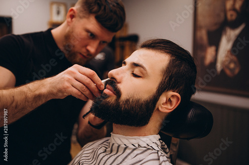 Getting perfect shape. Close-up side view of young stylish bearded man getting beard haircut by bearded, muscular hairdresser or barber at barbershop