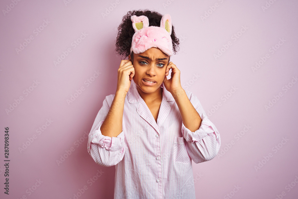Young african american woman wearing pajama and mask over isolated pink background covering ears with fingers with annoyed expression for the noise of loud music. Deaf concept. Young african american woman wearing pajama and mask over isolated pink background covering ears with fingers with annoyed expression for the noise of loud music. Deaf concept.