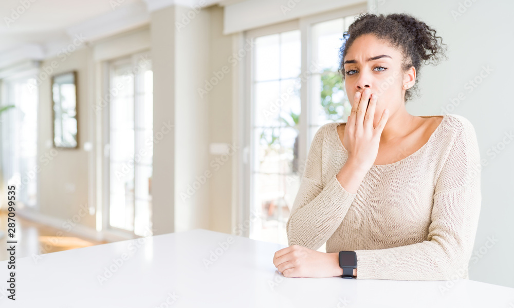 Beautiful young african american woman with afro hair sitting on table at home bored yawning tired covering mouth with hand. Restless and sleepiness. Beautiful young african american woman with afro hair sitting on table at home bored yawning tired covering mouth with hand. Restless and sleepiness.