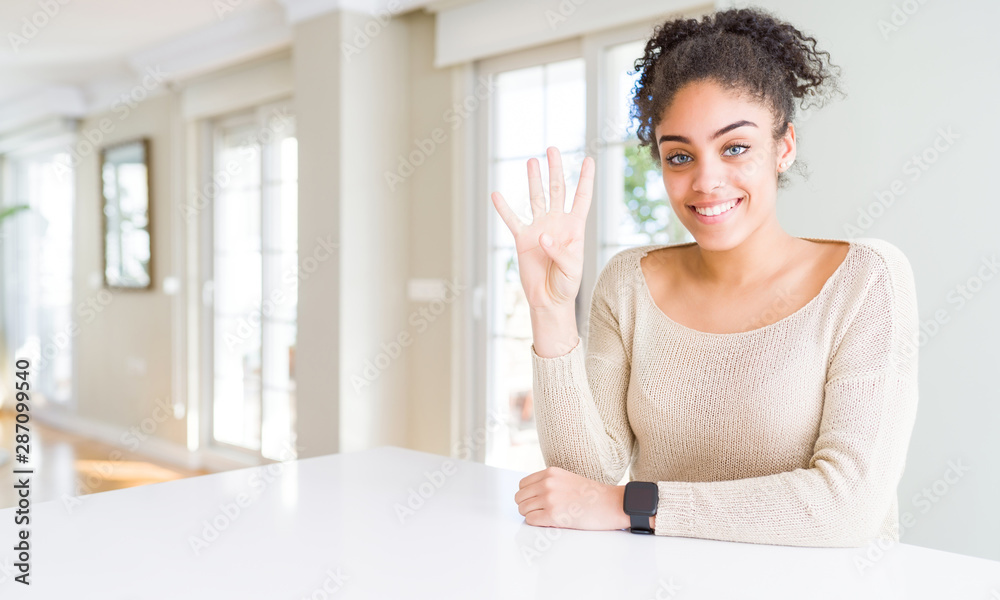 Beautiful young african american woman with afro hair sitting on table at home showing and pointing up with fingers number four while smiling confident and happy. Beautiful young african american woman with afro hair sitting on table at home showing and pointing up with fingers number four while smiling confident and happy.