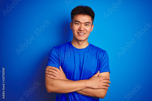 Young asian chinese man wearing t-shirt standing over isolated blue background happy face smiling with crossed arms looking at the camera. Positive person.