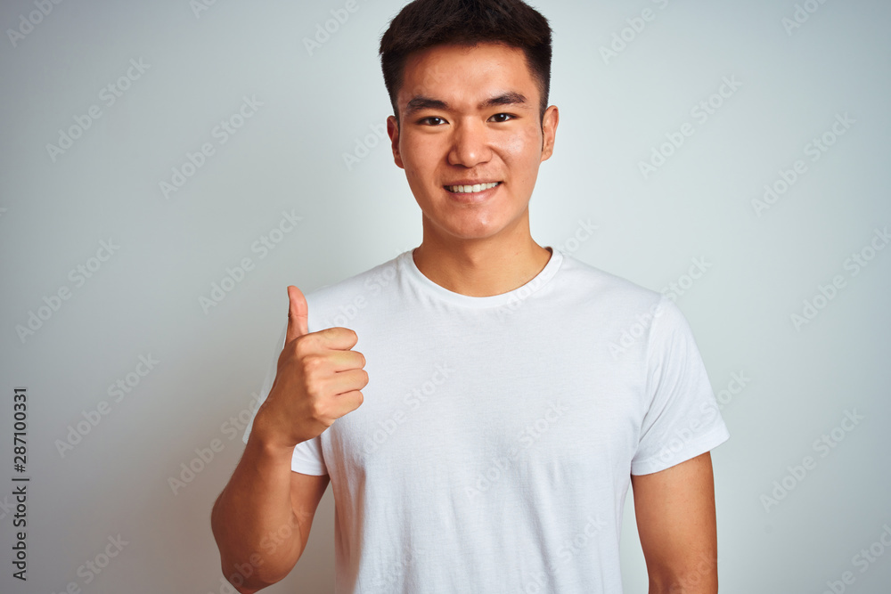 Young asian chinese man wearing t-shirt standing over isolated white background doing happy thumbs up gesture with hand. Approving expression looking at the camera showing success. Young asian chinese man wearing t-shirt standing over isolated white background doing happy thumbs up gesture with hand. Approving expression looking at the camera showing success.