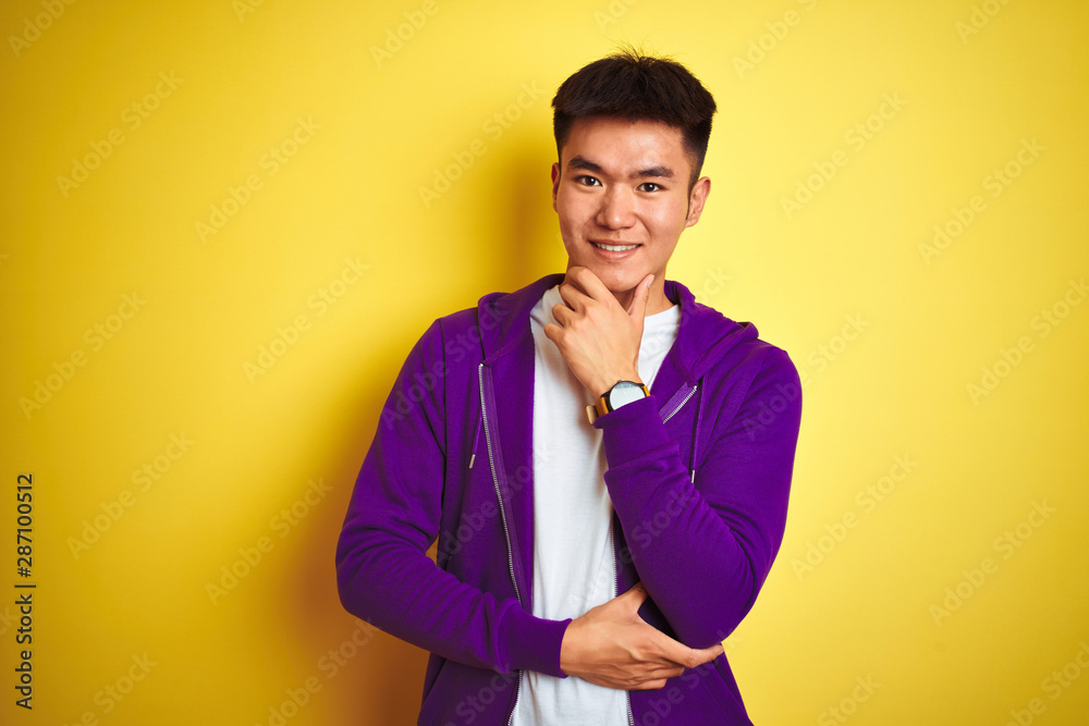 Young asian chinese man wearing purple sweatshirt standing over isolated yellow background looking confident at the camera smiling with crossed arms and hand raised on chin. Thinking positive.